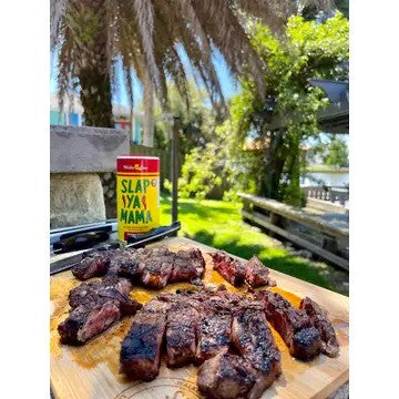 Grilled steaks on a cutting board with 'Slap Ya Mama' seasoning packet outdoors.